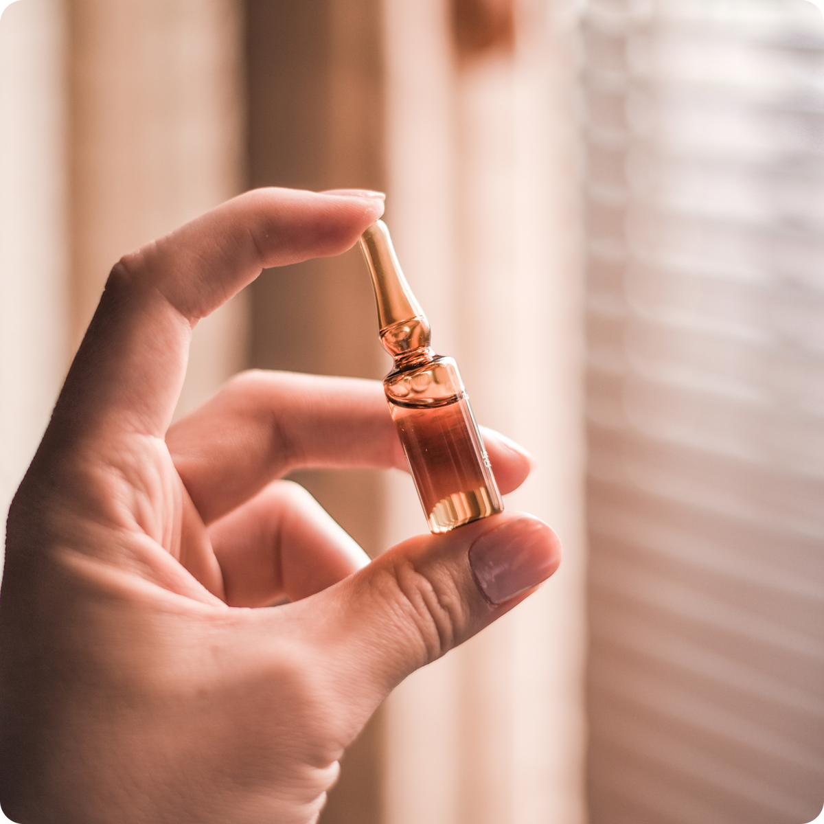 Close-up of hand holding amber glass serum ampoule for scalp treatment against a blurred window blinds background
