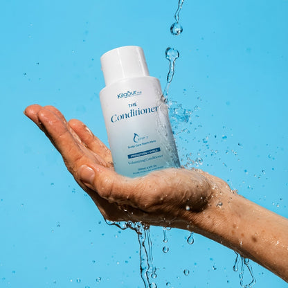 Hand holding a bottle of 'The Conditioner' with water droplets against a blue background