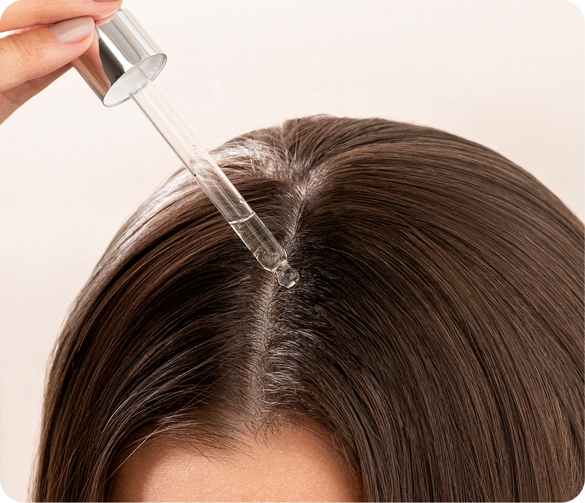 Close-up top view of scalp treatment serum dropper applying clear serum to center hair parting against a pale background