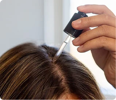 Person applying scalp serum with a dropper to the center part of brown hair